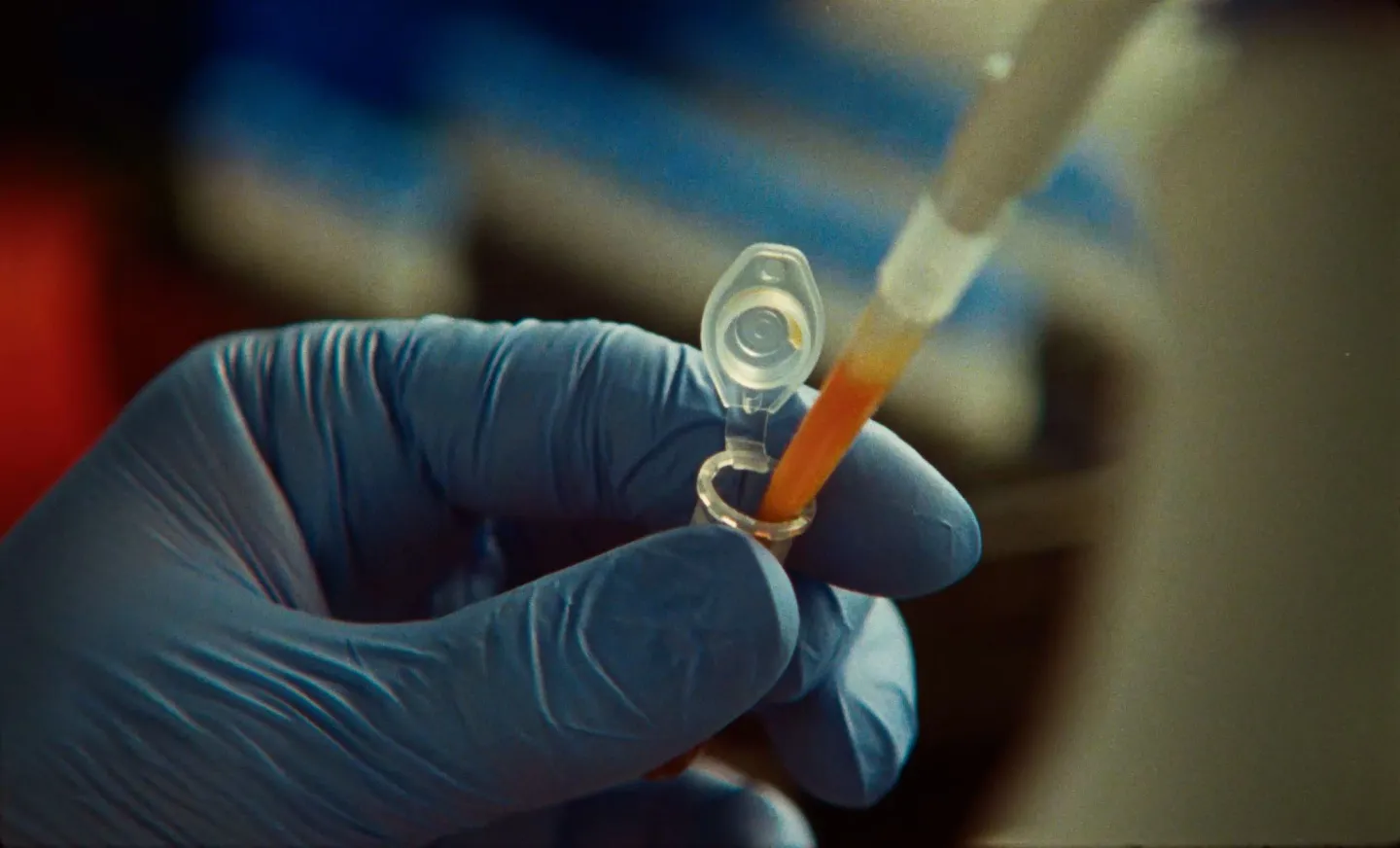 A hand in a blue lab glove holds an open plastic sample tube as a pipette tip filled with orange liquid reaches into it.