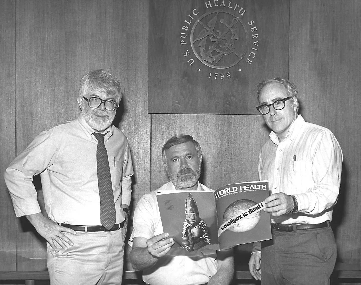 Drs. J. Donald Millar, William H. Foege and J. Michael Lane beneath the U.S. Public Health Service seal. Foege, seated, holds open the May 1980 issue of World Health, its cover declaring “smallpox is dead!”